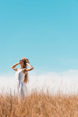 selective focus of stylish woman in white dress touching straw hat while standing with raised head against bule sky clipart