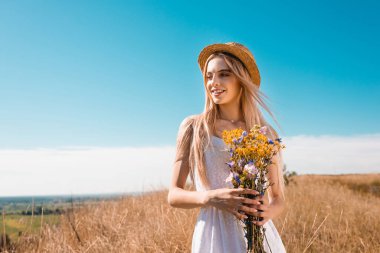 blonde woman in white dress and straw hat holding bouquet of wildflowers while looking away against blue sky clipart