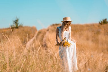 Beyaz elbiseli, saman şapkalı genç sarışın bir kadın elinde kır çiçekleriyle tarlada dikilirken...
