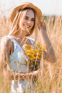 Çimen tarlasında kameraya bakarken kır çiçekleriyle hasır şapkaya dokunan genç sarışın kadının seçici odak noktası.