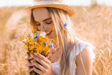 selective focus of blonde woman in straw hat smelling bouquet of wildflowers in grassy meadow clipart