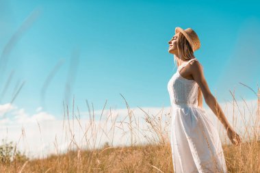 Beyaz elbiseli ve hasır şapkalı şık bir kadının seçici odak noktası uzanmış elleri ve kapalı gözleriyle çayırlarda duruyor.