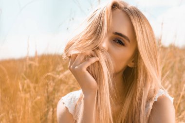 young blonde woman obscuring face with hair while looking at camera against blue sky in field clipart