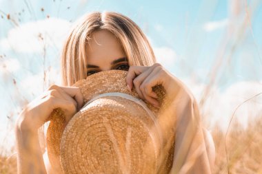 Mavi gökyüzüne doğru kameraya bakarken yüzü hasır şapkalı, seçici sarışın kadın odağı.