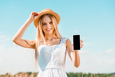 Beyaz elbiseli genç sarışın kadın hasır şapkaya dokunurken mavi gökyüzüne karşı beyaz ekranlı akıllı telefonu gösteriyor. 