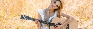 Hasır şapkalı sarışın kadının sahada akustik gitar çalarken kameraya bakışının yatay görüntüsü.