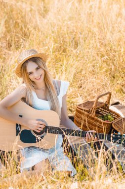 Hasır şapkalı sarışın kadının çayırda battaniyeyle akustik gitar çalarken kameraya baktığı seçici odak noktası.