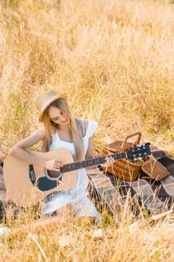 Beyaz elbiseli ve hasır şapkalı sarışının yüksek açılı görüntüsü. Battaniyede akustik gitar çalıyor. Seçici bir odak noktası.