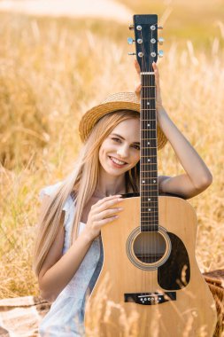 Çimenli alanda akustik gitarla otururken kameraya bakan genç sarışın kadının seçici odağı 