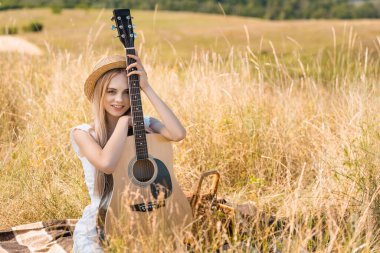 Sarışın kadın, yaz kıyafetleri ve hasır şapkalı akustik gitarla battaniyeye oturmuş kameraya bakıyor. 