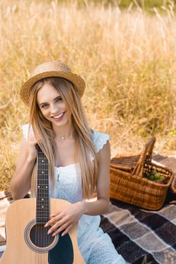 Beyaz elbiseli ve hasır şapkalı genç sarışın kadın hasır sepetin yanındaki battaniyede akustik gitarıyla oturuyor ve kameraya bakıyor.