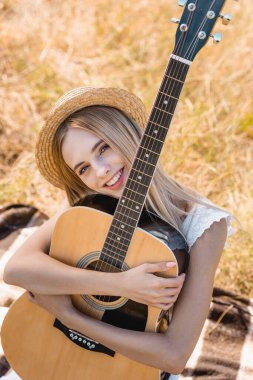 Hasır şapkalı sarışın bir kadının akustik gitarla battaniyede oturduğu yüksek açılı görüntüsü.