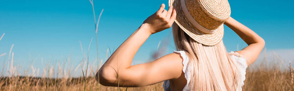 back view of blonde woman touching straw hat in grassy field against blue sky, horizontal image