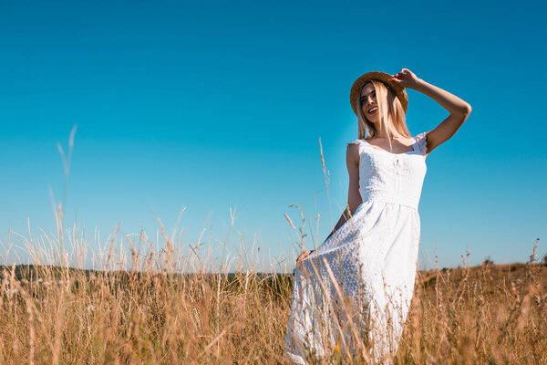 selective focus of stylish blonde woman touching straw hat and white dress while posing in grassy meadow against blue sky