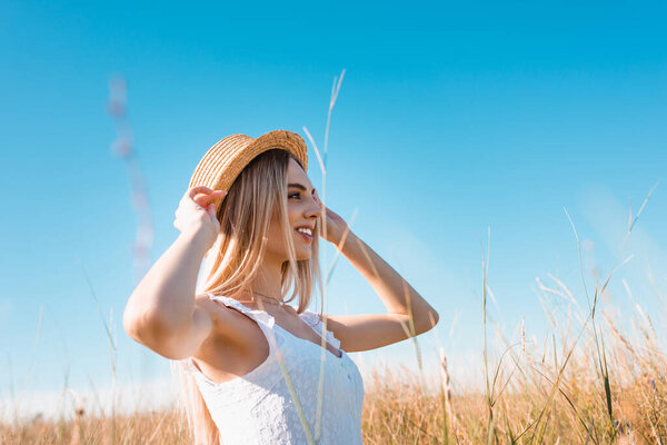 selective focus of stylish blonde woman touching straw hat while looking away against blue sky 