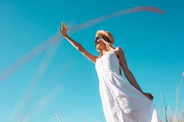 low angle view of stylish woman in white dress waving hand while looking away against blue sky, selective focus