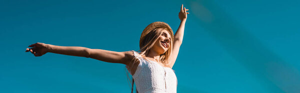horizontal image of young woman in straw hat standing with outstretched hands against blue sky, low angle view