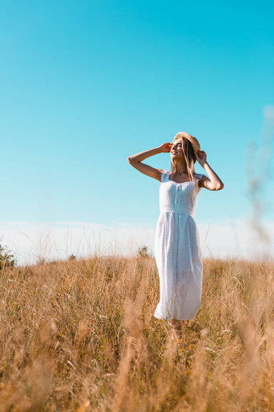 selective focus of blonde woman in white dress touching straw hat while standing in meadow against blue sky