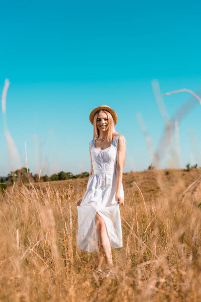 selective focus of sensual blonde woman in white dress and straw hat looking at camera while posing in grassland