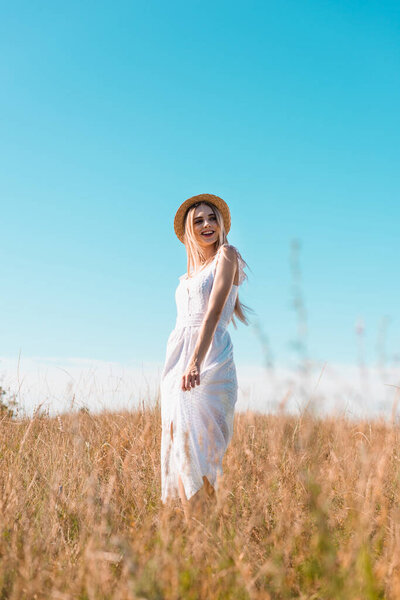 selective focus of blonde woman in white dress and straw hat standing in field against blue sky and looking away