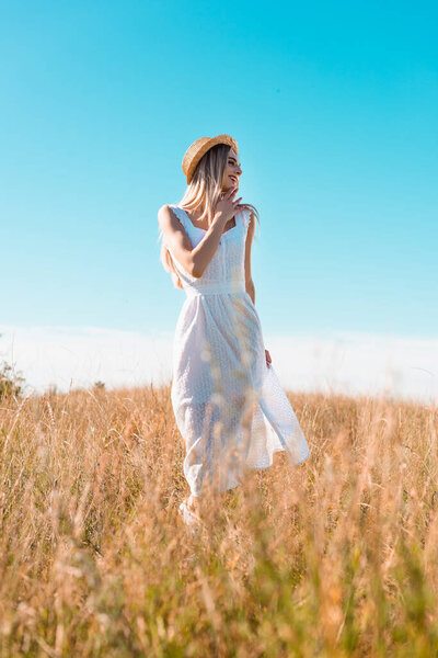 selective focus of stylish woman in white dress and straw hat touching chin and looking away in field against blue sky