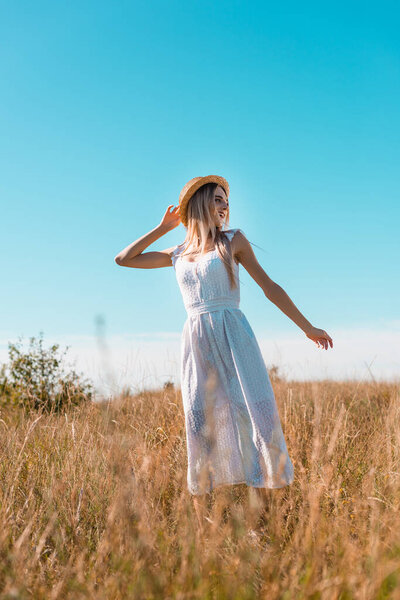 selective focus of blonde woman in white dress and straw hat showing follow me gesture in grassy meadow