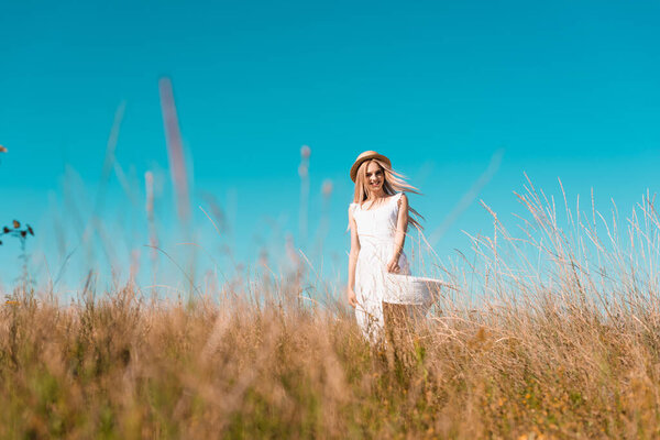 selective focus of stylish woman in white dress and straw hat looking at camera in grassy meadow against blue sky