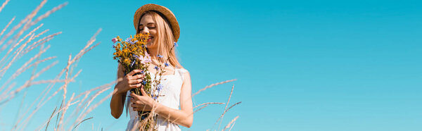 selective focus of young woman in straw hat holding bouquet of wildflowers against blue sky, panoramic concept