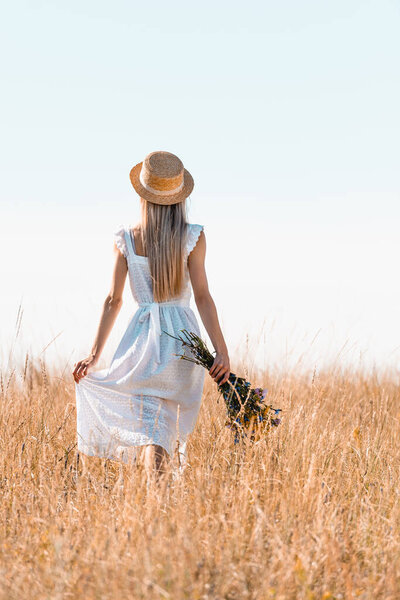 back view of woman in straw hat touching white dress while walking in grassy meadow with bouquet of wildflowers