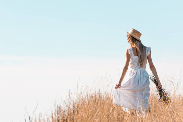 back view of young woman in straw hat touching white dress while holding wildflowers on grassy hill