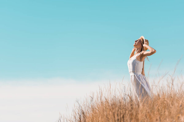selective focus of young woman in white dress touching straw hat while standing on grassy hill against blue sky