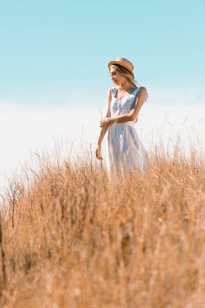 selective focus of young woman in white dress and straw hat looking away while standing on grassy hill