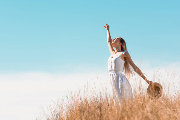 selective focus of blonde woman in white dress holding straw hat while standing with raised hand on grassy hill