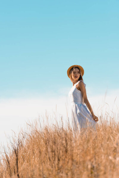 selective focus of young woman in straw hat touching white dress while standing on grassy hill