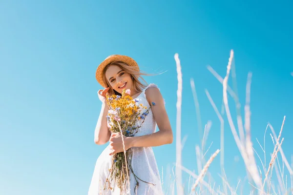 野の花を持ちながらカメラを見ながら髪に触れる白いドレスの女性の選択的な焦点 低角度ビュー — ストック写真