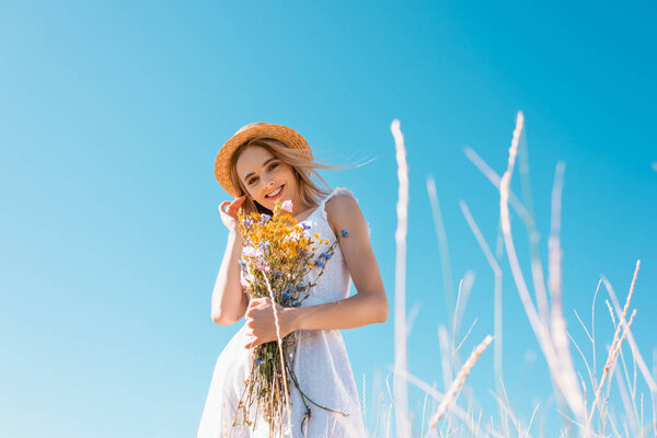 selective focus of woman in white dress touching hair while holding wildflowers and looking at camera, low angle view