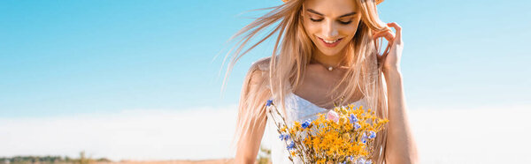 panoramic crop of sensual blonde woman holding bouquet of wildflowers against blue sky