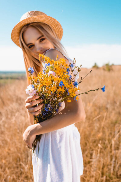 selective focus of blonde woman in straw hat holding wildflowers while looking at camera in grassy meadow