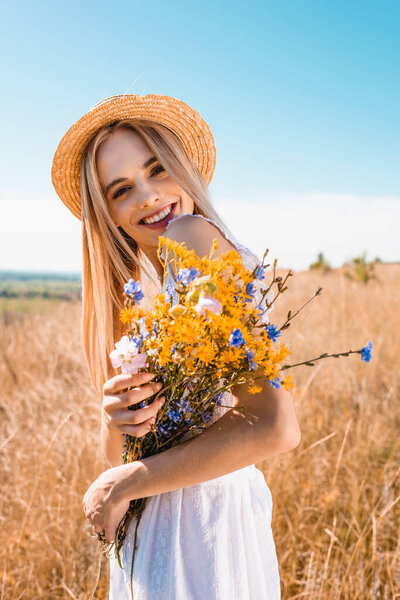 young stylish woman in straw hat holding wildflowers while looking at camera in grassy field