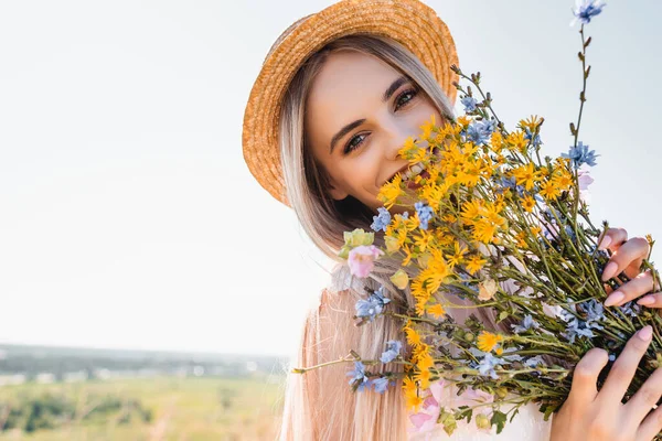Hasır Şapkalı Sarışın Kadın Elinde Kır Çiçekleri Tutarken Açık Gökyüzüne — Stok fotoğraf