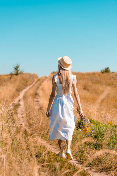 Вид сзади молодой женщины в белом платье и соломенной шляпе, идущей по полю с букетом полевых цветов