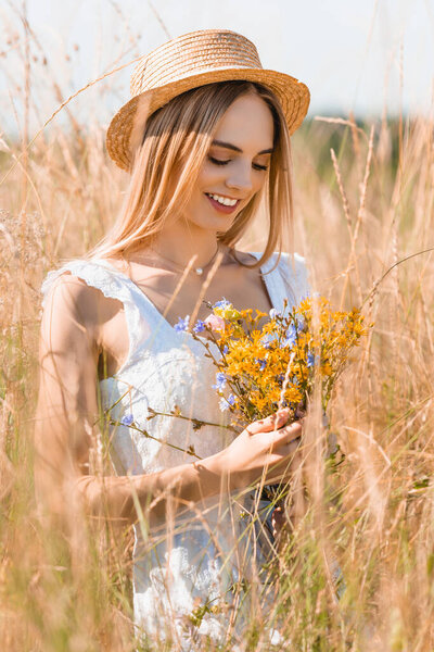 selective focus of stylish woman in white dress and straw hat holding wildflowers in grassy field