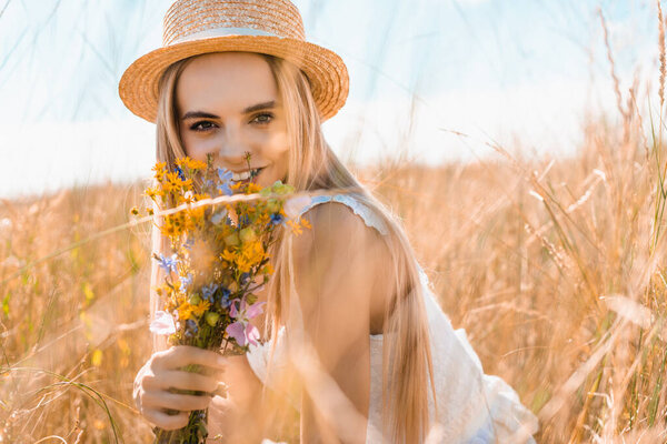 selective focus of young woman in straw hat holding wildflowers and looking at camera in grassy meadow