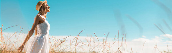 selective focus of young woman in white dress and straw hat standing with outstretched hands against blue sky, panoramic shot