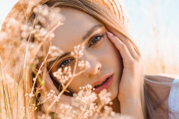 portrait of pensive blonde woman in straw hat touching face while looking at camera near wildflowers