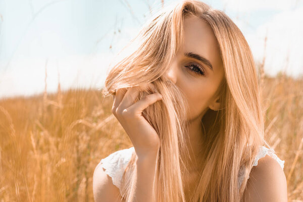 молодая блондинка затемняет лицо волосами, глядя в камеру на голубое небо в поле