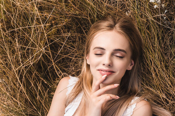 Top view of sensual blonde woman touching lips while lying on green grass with closed eyes