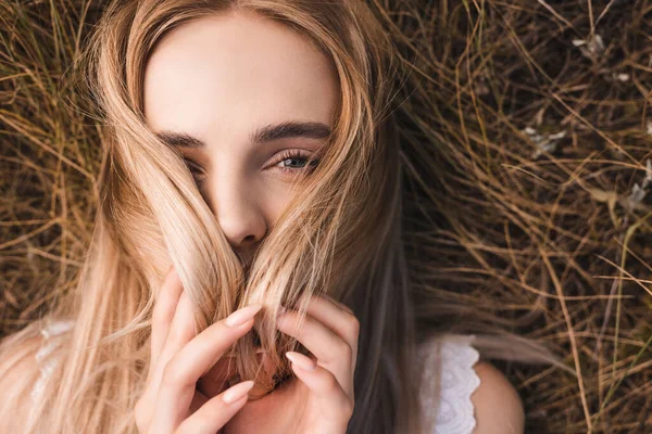 緑の草の上に横になり髪で顔を隠してカメラを見ている若いブロンドの女性のトップビュー — ストック写真