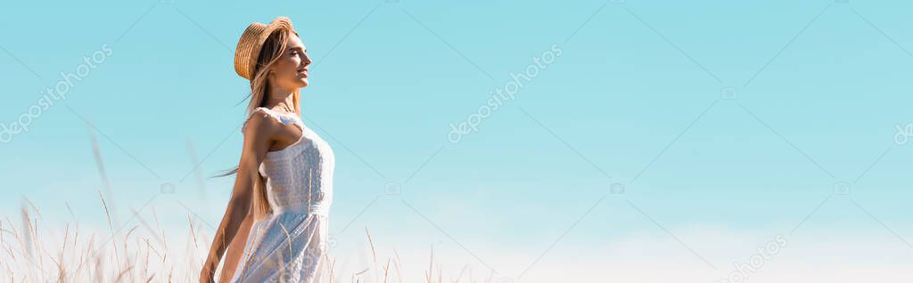 side view of young woman in white dress and straw hat standing against blue sky with outstretched hands, panoramic shot