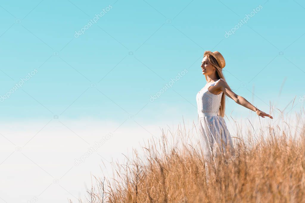 blonde woman in straw hat and white dress standing with outstretched hands on hill against blue sky